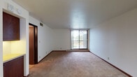 This single-floor residential unit offers a functional and comfortable layout designed to maximize livability. The main living area is generously sized, featuring neutral wall-to-wall carpeting, crisp white walls, and warm dark wood baseboards throughout. A sliding glass door opens to a private balcony, allowing natural light to fill the room and providing pleasant outdoor views of surrounding greenery. A ceiling fan adds practical comfort to the space.

The kitchen is thoughtfully equipped with rich medium-toned wood cabinetry offering ample storage, light-toned countertops, and a diagonal herringbone-patterned tile backsplash that adds visual interest. A white electric coil range serves as the central appliance, complemented by an overhead range hood.

The bedrooms are carpeted and feature large windows fitted with blinds or vertical shades, drawing in natural light while maintaining privacy. Baseboard heating units are present throughout, ensuring year-round comfort.

The bathroom includes a full tub and shower combination with floor-to-ceiling white subway tile accented by a decorative mosaic tile border. A standard toilet and tile flooring complete the clean, functional design. Overall, the unit presents a straightforward and well-maintained living environment suitable for comfortable everyday living.