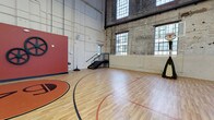 This sport court occupies a generously scaled single-floor space housed within a building of clear industrial heritage. The architecture is defined by dramatically high ceilings that emphasize the vertical volume of the room, lending the court an open, airy atmosphere well-suited for athletic activity. Exposed brick walls on one side retain the building's historic character, while whitewashed masonry walls on the opposing side provide a cleaner, more contemporary finish.

Large multi-pane industrial windows line multiple walls at both lower and upper levels, flooding the court with abundant natural light throughout the day. The polished hardwood floor is regulation-marked with clearly painted court lines in red and black, and features a prominent branded center-court logo in orange and black. A portable basketball hoop is positioned at one end of the court, while a bold red accent wall adorned with decorative industrial gear artwork serves as a striking visual focal point near the opposite baseline.

A black metal staircase leads to an upper-level access point, and traditional double doors with glass panes provide additional entry. Exposed overhead piping and industrial fixtures complete the utilitarian aesthetic, creating a dynamic and functional athletic environment that blends historic architectural character with modern sporting use.
