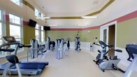 This fitness center offers a comprehensive workout environment designed to support a wide range of exercise routines. The space is outfitted with an array of strength training machines, including cable and resistance equipment, as well as cardio machines such as a treadmill and stationary bike, providing members with everything needed for a full-body workout.

The interior design is immediately eye-catching, with a bold color scheme featuring bands of olive green, coral red, and gold along the upper walls, creating a dynamic and motivating atmosphere. The light-colored, smooth flooring throughout the space provides a clean and functional surface, while large windows along one wall allow ample natural light to fill the room, enhancing the open and airy feel.

Overhead, modern linear pendant lighting ensures the space is well-lit at all times of day. A wall-mounted flat-screen television adds an entertainment option for those engaged in longer cardio sessions. The layout is thoughtfully arranged to allow comfortable movement between equipment. The overall ambiance is energetic yet welcoming, making this fitness center a well-rounded facility suited for both casual exercisers and dedicated fitness enthusiasts.