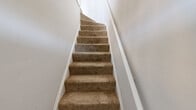This 2-bedroom, 1-bathroom unit offers a thoughtfully designed layout across a single floor, making efficient use of its compact footprint. The space is accessed via a carpeted staircase with crisp white walls, setting a clean and welcoming tone throughout.

The primary bedroom is a comfortable, well-proportioned room featuring plush beige carpeting, soft gray walls, and two windows fitted with blinds that allow natural light to filter in. A classic cast-iron radiator adds a charming architectural detail. The second bedroom features the same warm carpet flooring with white walls and direct access to a dedicated closet.

Common areas and the kitchen are finished with light hardwood flooring that lends warmth and continuity to the space. The kitchen is galley-style and features two-tone cabinetry — warm brown lower cabinets paired with white upper cabinets — complemented by a light marble-look countertop, a stainless steel sink, a gas range, and a distinctive blue subway tile backsplash that adds a modern pop of color.

The bathroom is a standout feature, showcasing floor-to-ceiling herringbone white tile in the tub surround, wood-look tile flooring, a clean white vanity with brushed nickel fixtures, and a window for natural ventilation. The space is further served by a dedicated closet, providing practical storage throughout.