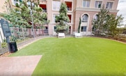 This meticulously designed outdoor space is a central feature of a multi-story residential complex in Irvine, California. The courtyard boasts a large, well-maintained artificial turf area, providing a lush green centerpiece that remains pristine year-round. The space is enclosed by decorative metal fencing, which offers both security and aesthetic appeal.

Strategically placed white benches provide comfortable seating areas for residents to relax and enjoy the outdoors. Tall pine trees and palm trees surround the courtyard, creating a natural backdrop and offering partial shade. The landscaping includes a variety of plants and shrubs, adding texture and color to the environment.

The building's architecture features a soft pink exterior with large, arched windows, creating an elegant backdrop for the courtyard. Outdoor lighting fixtures are installed throughout the space, ensuring the area is well-lit and inviting during evening hours.

This thoughtfully planned outdoor area serves as a communal gathering space, perfect for relaxation, social interactions, or quiet contemplation. It seamlessly blends natural elements with modern residential living, providing residents with a serene escape within their urban environment.
