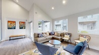 Located at 3202 W Bell Rd in Phoenix, AZ, this thoughtfully designed single-floor space serves as a welcoming community lounge, combining a living room and kitchen into one cohesive, functional layout. The living area immediately impresses with its dramatic vaulted ceilings that soar overhead, creating an expansive sense of openness that far exceeds the room's footprint. Crisp white upper walls transition into a bold slate-blue accent wall, adding depth and modern character to the space. Large windows along multiple walls flood the interior with abundant natural light and offer views of the surrounding outdoor grounds. Light wood-look plank flooring runs throughout, lending warmth and continuity. The living room is arranged for comfort and gathering, featuring a sectional sofa, swivel lounge chairs, a round coffee table, and a wall-mounted flat-screen TV. A patterned area rug in blue, gold, and ivory anchors the seating arrangement, while curated artwork completes the contemporary aesthetic. A sliding glass door provides convenient outdoor access. The compact kitchen is clean and efficient, outfitted with light wood cabinetry, white quartz countertops, a stainless steel sink, open display shelving, and an under-counter beverage refrigerator — ideal for casual entertaining and everyday convenience.