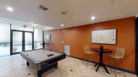 This well-appointed clubroom is a dynamic communal space designed to accommodate a variety of social activities. The layout is divided into distinct functional zones, each thoughtfully designed with its own character and purpose.

The lounge area is a warm and inviting space, anchored by comfortable sofas and armchairs arranged around a glass coffee table atop a colorful patchwork rug. Bold mustard-yellow walls and a striking sputnik-style ceiling fixture add a retro-modern flair, while floor-to-ceiling glass windows flood the room with natural light and offer views to the exterior.

Adjacent to the lounge, the fully equipped kitchen is finished in a sleek, contemporary style featuring dark cabinetry with gold hardware, dark granite countertops with subtle metallic flecks, a dark mosaic tile backsplash, and stainless steel appliances including a side-by-side refrigerator, range, and microwave. A large peninsula counter provides ample prep and serving space.

The game room area is defined by its deep terracotta-orange accent wall and light tile flooring. A full-size billiard table serves as the centerpiece, complemented by bistro-style high-top tables and woven rattan bar stools. Curated wall art throughout adds a polished, finished aesthetic to the overall space.