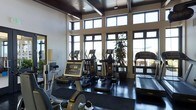 This fitness center is designed to deliver an exceptional workout experience within an architecturally refined setting. The space is defined by its impressive ceiling height, featuring dark-stained wood beam detailing that adds warmth and character overhead. A generous bank of large, multi-pane windows stretches across the exterior walls, reaching nearly to the ceiling and inviting abundant natural light throughout the day while offering expansive views of the surrounding landscape.

The main workout area is thoughtfully outfitted with a comprehensive selection of cardio equipment, including multiple elliptical trainers, treadmills, and a stationary cycle, all arranged to take full advantage of the scenic views outside. The flooring features durable, dark rubberized matting suited for high-traffic fitness use. Lush indoor plants are incorporated into the layout, contributing a fresh, welcoming atmosphere to the environment.

The combination of soaring ceilings, natural light, and modern equipment creates a bright and motivating space. The overall ambiance strikes a balance between functional athletic facility and upscale design, making it a standout amenity that encourages regular use and an active lifestyle.