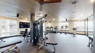 This fitness center in Dallas, TX, is designed to support a comprehensive workout experience within a thoughtfully appointed single-floor space. The main exercise area features a distinctive wood-planked ceiling with modern ceiling fans, recessed lighting, and large windows that invite natural light and outdoor views, creating an open and energizing atmosphere. Durable, neutral-toned carpet flooring spans the space, providing comfort underfoot across all workout zones.

The cardio section is equipped with multiple treadmills and elliptical machines, each fitted with individual screens, as well as stationary cycling bikes positioned along the windowed wall. A dedicated strength training zone houses a multi-station cable machine, free weights on tiered dumbbell racks, and weight benches. Wall-mounted flat-screen televisions are positioned throughout for entertainment during workouts.

A separate functional training room offers a more intimate setting, featuring textured dark accent walls, floor-to-ceiling mirrors, a ceiling fan, and a wall-mounted rack with TRX suspension trainers, kettlebells, and a medicine ball. This versatile room is ideal for stretching, bodyweight training, or guided workouts. Decorative touches such as framed artwork and accent decor contribute to an upscale, modern aesthetic throughout the facility.