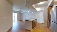 This single-floor space is designed with an open-concept layout that integrates the kitchen and living areas into one cohesive, functional environment. The kitchen is anchored by a substantial central island finished with a dark countertop, an undermount sink with a modern faucet, and warm wood cabinetry with brushed metal hardware. Two pendant lights hang above the island, providing focused task lighting that complements the overall design. A notable skylight above the kitchen area introduces an abundance of natural light, brightening the space throughout the day. The flooring throughout is hardwood-style, lending warmth and continuity across the open plan. The living area extends seamlessly from the kitchen, featuring a ceiling fan and access to an outdoor balcony through large sliding glass doors, which allow additional natural light and views of the exterior. The neutral color palette of soft grey walls and white trim creates a clean, versatile backdrop. A paneled door on the far wall leads to additional areas of the unit, suggesting further private spaces beyond the main open living zone.