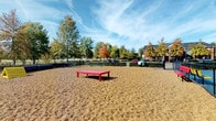 Bark Park is a well-appointed outdoor dog park designed to provide a safe and engaging environment for dogs and their owners. The park is fully enclosed by a black chain-link fence, with dense shrub hedging along portions of the perimeter that adds a natural buffer from the surrounding area. The ground throughout the main play area is covered in a generous layer of pea gravel, offering a comfortable, low-maintenance surface for dogs to run and explore. Scattered throughout the space are colorful amenities, including a bold red elevated platform for dogs to climb and a yellow agility tunnel, providing opportunities for interactive play. Vibrant benches in red and yellow offer seating for owners while they supervise their pets. Adjacent to the main gravel area lies a vividly painted multi-sport court surfaced in bright blue with colorful markings, separated by the chain-link fencing and accessible via a gated entry. Mature deciduous trees line the perimeter, providing seasonal shade and a pleasant natural backdrop under open skies. The park is situated within a residential community, with brick buildings and additional greenery visible beyond the fence, creating a neighborhood-friendly setting that is both functional and inviting for daily use.