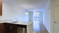 This single-floor space showcases an open-concept layout that seamlessly integrates the kitchen and living area into one cohesive environment. The kitchen features a peninsula with a light stone countertop, an undermount sink with a modern faucet, dark cabinetry, and a stainless steel dishwasher. A pendant light hangs above the peninsula, providing focused task lighting while contributing to the space's contemporary aesthetic. Crown molding lines the ceilings throughout, adding a refined architectural detail. The living area extends generously from the kitchen, featuring warm hardwood flooring that runs throughout the space, enhancing the sense of continuity. A ceiling fan is centrally positioned in the living area, offering both function and subtle style. Natural light floods the interior through a large floor-to-ceiling window and glass door at the far end of the living area, which opens to what appears to be a balcony with an exterior railing. The bright, neutral white walls amplify the natural light, creating an airy and welcoming atmosphere. Overall, the space presents a clean, modern aesthetic with practical features and an abundance of natural light.