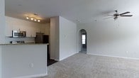 This single-floor residence offers a thoughtfully designed layout that balances open living spaces with defined private areas. The kitchen serves as a central hub, featuring granite-look countertops, white cabinetry, stainless steel appliances including a refrigerator, and a peninsula that separates it from the living area. Dark hardwood-style flooring grounds the kitchen zone, while the adjoining living area transitions to plush carpeting, creating a warm and comfortable atmosphere.

A distinctive arched doorway connects the main living space to a secondary carpeted room, adding an architectural detail that enhances the overall character of the home. Built-in shelving within the arched nook provides convenient storage and display options. The open living and dining area is generously proportioned, allowing for flexible furniture arrangements.

The floor plan, as viewed from above, reveals multiple rooms including bedrooms and bathrooms arranged along the perimeter, each benefiting from exterior windows that allow natural light to filter throughout. The neutral color palette of soft grays and whites across the walls and ceilings creates a clean, cohesive aesthetic that flows seamlessly from room to room.