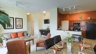 This single-floor residence showcases an open-concept layout that seamlessly connects the dining area, living room, and kitchen into one cohesive living space. The dining area is anchored by a granite-topped counter or table, complemented by dark leather and wood-frame chairs that add a touch of sophistication. Adjacent to the dining space, the living room offers a comfortable seating arrangement with neutral-toned sofas accented by warm orange throw pillows, centered around a stone-topped coffee table. A wall-mounted flat-screen television serves as a focal point along the main wall. The kitchen is distinguished by a bold burnt-orange accent wall that creates visual warmth, paired with maple-toned cabinetry, dark countertops, and stainless steel appliances. Track lighting overhead ensures the kitchen is well-lit and functional. Light-colored carpeting flows through the living areas, contributing to a bright and airy ambiance. Neutral white walls throughout most of the space allow the warm accent colors and rich furnishings to stand out. The open floor plan creates an inviting, connected atmosphere ideal for both everyday living and entertaining.