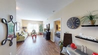 Located at 7027 Seguin Rd in San Antonio, Texas, this single-floor residence offers a thoughtfully arranged layout that balances comfort and functionality. The open living area is bathed in natural light from large windows dressed with sheer curtains, creating a bright and welcoming atmosphere. Rich wood-style flooring flows throughout the main living spaces, adding warmth and visual continuity.

The living room provides ample space for seating and entertaining, featuring neutral-toned walls that lend a calm, versatile backdrop. Adjacent to the living area, the kitchen is equipped with warm oak cabinetry extending to the upper walls, granite-style countertops, and a full suite of black appliances including a refrigerator, range, dishwasher, and microwave. A dining area sits just off the kitchen, completing the open common area.

The bedroom is a comfortable retreat with plush carpeting, a ceiling fan, and a window that allows natural light to filter in. It connects directly to a full bathroom outfitted with a combination tub and shower, wood-style flooring, and a vanity with under-sink storage. A spacious walk-in closet with built-in wire shelving provides generous storage. An additional utility closet with wire shelving offers even more organizational space, rounding out this practical and comfortable San Antonio residence.