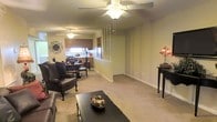 This single-floor space features an open-concept layout that seamlessly integrates the living, dining, and kitchen areas into one cohesive environment. The living area is carpeted in a neutral beige tone that extends throughout the space, creating a warm and unified aesthetic. A ceiling fan with a light fixture is centrally positioned, providing both ventilation and ambient lighting to the main living zone.

A decorative spindle partition subtly delineates the living area from the rest of the space without fully closing it off, maintaining the open feel. The dining area accommodates a table with seating and is positioned adjacent to the kitchen, which features standard cabinetry, a stainless steel refrigerator, and a window that allows natural light to filter in.

The walls are finished in a soft, warm beige, complementing the carpeted floors and contributing to the overall cozy ambiance. Decorative elements, including wall art and accent lighting, add character to the space. White baseboards and trim provide clean contrast throughout. The layout is practical and functional, offering a comfortable flow between all primary living zones on a single level.