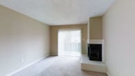 This single-floor space offers a clean and functional layout with a warm, neutral aesthetic throughout. The main living area is generously proportioned, featuring plush wall-to-wall carpeting in a light beige tone that complements the soft, neutral-painted walls. A standout feature of the living area is the corner fireplace, clad in classic white tile with a raised tile hearth and a black firebox, providing a cozy focal point that adds both character and warmth to the room. Natural light pours into the space through a large sliding glass door fitted with vertical blinds, which opens onto an outdoor balcony, offering a seamless connection to the exterior. The textured popcorn ceiling and white baseboards complete the traditional interior finish of the space. The overall ambiance is calm and understated, providing a neutral canvas well-suited for a variety of uses and personal styles. The single-floor layout ensures easy accessibility and a straightforward flow throughout the space.