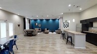 This well-appointed single-floor space showcases a thoughtfully designed open-concept layout that seamlessly connects the living and kitchen areas. The living room serves as the centerpiece, anchored by a striking teal accent wall that adds a bold pop of color against the neutral grey tones throughout. Decorative mirrors adorn the accent wall, complementing the contemporary furnishings that include grey upholstered sofas, a glass-and-wood coffee table, and coordinating side tables. Recessed lighting illuminates the space, while navy blue curtains frame the windows, allowing for natural light and adding a cohesive design element.

The kitchen is efficiently designed with dark espresso cabinetry contrasting sharply against light granite countertops. Stainless steel appliances — including a refrigerator, range, and microwave — provide full cooking functionality. A peninsula counter with bar seating, highlighted by elegant pendant lighting, bridges the kitchen and living area, making it ideal for casual dining or entertaining.

Light wood-look flooring runs consistently throughout the entire space, contributing to a clean, modern aesthetic. The layout offers generous open floor space, creating an airy and functional environment suitable for both relaxation and social gatherings.