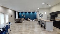 This well-appointed single-floor space showcases a thoughtfully designed open-concept layout that seamlessly connects the living and kitchen areas. The living room serves as the centerpiece, anchored by a striking teal accent wall that adds a bold pop of color against the neutral grey tones throughout. Decorative mirrors adorn the accent wall, complementing the contemporary furnishings that include grey upholstered sofas, a glass-and-wood coffee table, and coordinating side tables. Recessed lighting illuminates the space, while navy blue curtains frame the windows, allowing for natural light and adding a cohesive design element.

The kitchen is efficiently designed with dark espresso cabinetry contrasting sharply against light granite countertops. Stainless steel appliances — including a refrigerator, range, and microwave — provide full cooking functionality. A peninsula counter with bar seating, highlighted by elegant pendant lighting, bridges the kitchen and living area, making it ideal for casual dining or entertaining.

Light wood-look flooring runs consistently throughout the entire space, contributing to a clean, modern aesthetic. The layout offers generous open floor space, creating an airy and functional environment suitable for both relaxation and social gatherings.