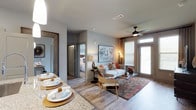This single-floor residence showcases a thoughtfully designed open-concept layout that connects the dining and living spaces in a seamless, functional flow. The kitchen peninsula features striking granite countertops and is illuminated by elegant pendant lights, making it a natural gathering point. The dining area is set beneath the warm glow of pendant lighting, offering a polished yet comfortable setting for meals and entertaining.

The living area is anchored by a contemporary ceiling fan with a sleek dark finish, complementing the neutral grey walls and light hardwood flooring that run throughout the space. A stylishly arranged gallery wall adds a personalized, curated touch above the sofa, which is adorned with coordinating accent pillows in muted tones and a pop of mustard yellow. Natural light filters into the room through a large window dressed with layered curtains, enhancing the bright and airy ambiance.

The overall aesthetic is modern and cohesive, blending clean lines with warm textures and metallic accents. The space feels open and well-proportioned, offering a comfortable and visually appealing environment suited for both everyday living and entertaining.