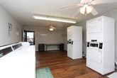 This single-floor shared laundry facility is designed for practical, communal use, offering a well-organized and functional layout. The main washing area features a row of top-load washing machines lined along one wall, providing ample capacity for multiple users. Adjacent to the washers, a dedicated drying room houses several stacked dryer units, maximizing floor space efficiency while accommodating high-volume laundry needs. Instructional signage is posted throughout to guide users on proper machine operation.

The space is grounded by warm hardwood flooring that runs throughout, lending a clean and welcoming feel to what is typically a utilitarian environment. A ceiling fan with a light fixture provides air circulation and overhead lighting in the main area, keeping the space comfortable during use. A folding table positioned near the entrance offers a convenient surface for sorting and folding laundry, while wall-mounted hooks provide additional organization.

A row of seating along one wall offers a comfortable waiting area for patrons. The facility is well-lit and features clearly posted no-smoking and security notices. A glass-paneled entry door allows natural light to filter in, connecting the interior to the surrounding exterior. Overall, the space is clean, efficiently arranged, and purpose-built to serve the laundry needs of a shared community.