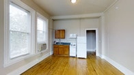 This single-floor space is characterized by its consistent design language of warm hardwood flooring, neutral beige and gray walls, and crisp white trim and door frames that run throughout. A central hallway connects the various rooms, offering an open and navigable layout with generous doorway openings that enhance the sense of flow between spaces. Natural light filters in through multiple windows, brightening the interior and highlighting the natural tones of the wood floors.

The kitchen area is compact yet functional, equipped with warm wood cabinetry, a dark countertop with an integrated sink, a white range, a full-size refrigerator, and a microwave. A window unit provides climate control, and large windows allow ample natural light into the space. The kitchen opens directly into an adjoining room, maintaining a connected layout.

The remaining rooms continue the same palette of light walls and hardwood floors, with white-painted built-in storage elements visible in one of the rooms. The doorways are framed with substantial white molding that adds a classic architectural touch. Overall, the space presents a clean, straightforward interior with functional features and consistent finishes throughout.