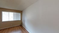 This single-story space presents a straightforward, well-maintained interior with a neutral color palette that provides a versatile backdrop for a variety of uses. The room showcases warm-toned, wood-look hardwood flooring that adds character and is easy to maintain. Walls are finished in a clean off-white tone, complemented by tan-colored accent walls that introduce subtle warmth to the overall aesthetic. A textured popcorn ceiling adds a classic touch overhead, while white baseboards provide a clean transition between the walls and flooring. A single window fitted with horizontal blinds allows natural light to filter into the space, creating a bright and airy atmosphere while offering privacy control. The room's simple, open layout provides flexibility for various furniture arrangements or functional configurations. The overall ambiance is clean, understated, and move-in ready, making it a practical and adaptable space suited for everyday living or use.