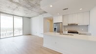 This contemporary single-floor unit showcases a thoughtful blend of industrial and modern design elements throughout. Exposed concrete ceilings contrast elegantly with crisp white walls and light wood-tone flooring, creating a clean yet characterful aesthetic across all rooms.

The open-concept kitchen serves as a central hub, featuring a large waterfall-edge island with an undermount sink, sleek flat-front white cabinetry extending to the ceiling, subway tile backsplash, and a full suite of stainless steel appliances including a French door refrigerator and range. Recessed lighting keeps the space bright and functional.

The living area flows naturally from the kitchen and benefits from floor-to-ceiling sliding glass doors that open to an outdoor balcony, flooding the space with natural light and offering expansive elevated views of the surrounding cityscape and greenery.

Each bedroom is generously proportioned, featuring the same light hardwood flooring and exposed concrete ceilings. Several bedrooms include their own sliding glass door or large window access to outdoor views, enhancing the sense of openness.

Two well-designed bathrooms complete the unit. The primary bath includes a glass-enclosed walk-in shower with white subway tile walls, a bench, mosaic patterned floor tile, and a floating vanity with globe sconce lighting. The second bathroom mirrors the refined aesthetic with a soaking tub, subway tile surround, and a matching decorative floor tile.