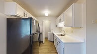 This well-organized 2-bedroom, 1-bathroom residence is laid out entirely on a single floor, making efficient use of its compact footprint. The open-concept kitchen and living area forms the heart of the home, connected by wood-look laminate flooring throughout. A structural column divides the two spaces while maintaining visual openness, with a sliding glass door in the living area inviting natural light and providing direct access to the outdoors and greenery beyond.

The galley-style kitchen is thoughtfully designed with white cabinetry featuring black hardware, white countertops, a single-basin sink, and black appliances including a full-sized refrigerator. A dedicated laundry area is accessible directly from the kitchen, adding everyday convenience. Baseboard heating units are present throughout the living spaces.

Both bedrooms are carpeted and offer a quiet, neutral atmosphere. The primary bedroom includes a sliding closet and benefits from natural light, while the second bedroom features a window with an outdoor view. The full bathroom is clean and functional, equipped with a tub-shower combination. Textured ceilings are consistent throughout the unit, contributing to the home's cohesive, understated character.