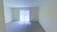 This single-story space features a spacious main living area finished with wall-to-wall carpeting in a neutral tone, providing a comfortable and understated foundation. The room is painted in a soft, light yellow hue that contributes to an open and airy atmosphere. A prominent sliding glass door serves as the focal point of the space, framed by full-length vertical blinds that filter and diffuse natural light generously throughout the room. When the blinds are open, sunlight streams across the carpet in dramatic lines, enhancing the brightness of the interior. The textured white ceiling adds a subtle architectural detail overhead, while clean white baseboards run along the perimeter of the room, offering a crisp contrast to the neutral walls and flooring. The layout is open and unobstructed, lending itself well to flexible furniture arrangements. The sliding glass door likely provides access to an outdoor area, adding potential for indoor-outdoor connectivity. Overall, the space presents a clean, functional, and well-lit environment suitable for a variety of uses.