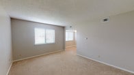 This single-floor unit offers a straightforward, functional layout across several well-defined spaces. The living area is generously sized, finished with neutral-toned wall-to-wall carpeting and soft gray walls. Multiple windows fitted with horizontal blinds allow natural light to filter throughout, creating a bright and open atmosphere. A textured popcorn ceiling runs consistently through the main living spaces, adding a classic residential character.

The floor plan, visible in the 3D model, reveals a connected layout that flows from the living area through to additional rooms, with tile flooring transitioning into carpeted zones. A dedicated bedroom includes a ceiling fan with light fixture, a closet, and a window overlooking the exterior — offering both ventilation and natural light.

The kitchen is equipped with warm-toned wood cabinetry, granite-style countertops, and a white gas range, providing a practical cooking space. Adjacent to the kitchen, the bathroom features full white square tile surrounding a soaking tub with an overhead shower, chrome fixtures, and light tile flooring — delivering a clean, utilitarian design. Throughout the unit, white baseboards and neutral finishes contribute to a cohesive, move-in-ready aesthetic.