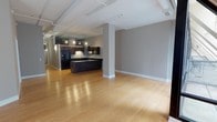 This single-floor loft-style unit showcases a compelling blend of industrial character and modern refinement. The expansive open-concept living area is anchored by warm hardwood flooring that reflects light throughout, while exposed white piping runs along the ceiling, lending an authentic loft aesthetic. The kitchen is a standout feature, equipped with rich dark espresso cabinetry, sleek black granite countertops, a central island, and black stainless appliances including a range and built-in microwave. Track lighting overhead keeps the space bright and functional. The bedroom areas are finished with plush neutral carpet and feature oversized black-framed floor-to-ceiling windows that flood the rooms with natural light and frame views of the surrounding urban cityscape. The unit includes two bathrooms: one with a full soaking tub and dark slate-style tile surround paired with a dark wood vanity and granite countertop, and a second with a glass-enclosed walk-in shower finished in matching dark tile. The overall palette of warm grays, deep espresso tones, and light wood flooring creates a cohesive, contemporary ambiance throughout this well-appointed urban residence.
