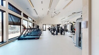 This expansive, single-level fitness center is designed with both functionality and aesthetics in mind. The space features soaring vaulted ceilings equipped with sleek linear pendant lighting and multiple ceiling fans, creating an open, airy atmosphere throughout. Large floor-to-ceiling windows line the walls, flooding the interior with natural light and offering direct views of the surrounding outdoor pool and courtyard areas.

The main workout floor is finished with a smooth, light-colored resilient flooring that spans the entire space. The cardio zone is well-appointed with a row of professional-grade treadmills and elliptical cross-trainers positioned along the windowed wall, allowing users to exercise while enjoying outdoor views. A dedicated strength training area houses a comprehensive selection of cable machines, multi-functional weight stations, a dumbbell rack, and various resistance equipment.

A smaller, comfortable lounge area features built-in bench seating with colorful accent cushions and an interior window overlooking the main gym floor, providing a relaxed space for rest or observation. The facility also includes package lockers near the entrance. The overall design balances a clean, modern aesthetic with a welcoming, well-lit environment suited for a full range of fitness activities.