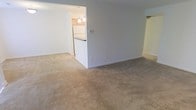 This single-floor residential unit offers a straightforward and functional layout designed for comfortable everyday living. The main living area is generously sized, featuring soft neutral carpeting that extends throughout, clean white walls, and standard baseboards that give the space a simple, move-in-ready feel. The room benefits from a calm, understated ambiance with ample space for a variety of furniture arrangements.

A distinctive pass-through opening connects the living area directly to the kitchen, creating a sense of openness between the two spaces. The kitchen is equipped with warm wood-toned cabinetry, a white refrigerator, and a peninsula-style breakfast bar that serves as both a prep and dining surface. Overhead, pendant lights hang from the ceiling, providing focused task lighting and adding a touch of character to the kitchen area.

Natural light filters into the space, keeping the interiors bright and welcoming. The overall design is clean and practical, with a neutral color palette throughout that provides a versatile backdrop for personalization. The unit's single-level layout ensures easy accessibility and a cohesive flow from one area to the next, making it a practical and livable space.