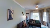 This single-floor space offers a thoughtfully designed interior that balances comfort and style. The living room serves as the centerpiece, showcasing elegant crown molding that frames the ceiling and adds a refined architectural touch. Soft, neutral wall tones — including a calming mint-green accent wall — create a welcoming and airy atmosphere throughout the space.

Large windows dressed with patterned curtains allow generous amounts of natural light to flood the room, offering pleasant views of the surrounding greenery. A ceiling fan provides both functional airflow and a subtle decorative element. The living area is anchored by plush dark sofas accented with colorful throw pillows, arranged around a geometric-patterned area rug that adds visual interest to the carpeted floor.

A dedicated media area features a television atop a dark wood entertainment console, while a coordinating sideboard provides additional storage and display space. Wall art and decorative accessories are carefully curated to complement the overall aesthetic. A full-length mirror near the entryway enhances the sense of space. The layout flows naturally between areas, making the space feel open and well-connected. Overall, the space exudes a modern yet cozy ambiance suited for everyday living.