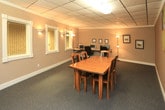 Located at 3901 Indian School Rd NE in Albuquerque, New Mexico, this single-floor commercial space offers a practical and well-appointed environment suited for professional use. The interior is finished with wall-to-wall gray carpeting and clean white baseboards that run throughout, lending a cohesive and polished look to the space.

The main area serves as a versatile room combining conference and workstation functions. A warm-toned wooden conference table with matching chairs anchors one section of the room, while a dedicated workstation area along the wall accommodates multiple computer setups. Drop-panel ceiling tiles with integrated fluorescent lighting and supplemental floor lamps provide consistent, functional illumination throughout.

The walls are finished in a warm taupe tone with textured surfaces, creating a neutral and professional atmosphere. Framed artwork and photographs add modest decorative touches. Interior windows with white trim and wire-glass panes contribute to the open feel of the space while maintaining a clean, commercial aesthetic.

Entry is through a white door with a gold push bar and wire-glass window, reflecting standard commercial accessibility standards. A clearly marked exit sign is visible, confirming compliance with safety requirements. Overall, this space presents a functional, move-in-ready environment ideal for office, administrative, or collaborative professional use.