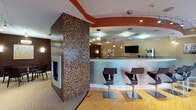 Located at 399 Main Ave in Norwalk, Connecticut, this single-floor amenity space is designed with a sophisticated, contemporary aesthetic that balances comfort and functionality. The centerpiece of the space is a dramatic floor-to-ceiling mosaic tile column housing a sleek, modern fireplace, its warm earth-toned tiles creating a striking focal point that anchors the open layout.

The space transitions fluidly between distinct functional zones. A curved bar counter with stylish high-back swivel stools provides a social gathering area, backed by a wall-mounted flat-screen TV and pendant lighting that adds a refined ambiance. Adjacent to the bar, a fully equipped kitchenette features granite countertops, mosaic tile backsplash, warm wood cabinetry, a microwave, beverage cooler, and a water and coffee station — ideal for hosting events or everyday use.

A dedicated lounge area offers plush wicker seating with accent pillows arranged around a second flat-screen television set against a rich, dark accent wall. A separate dining nook with a round table and chairs provides a more intimate setting beneath elegant pendant lighting. The entry area welcomes visitors with hardwood flooring, glass storefront walls allowing natural light, and a comfortable leather seating arrangement. Track and pendant lighting throughout enhances the warm, inviting atmosphere of the entire space.