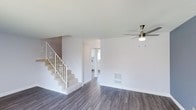 This two-floor home offers a clean, functional layout designed for comfortable everyday living. The main level showcases a spacious open living area finished with grey hardwood-style flooring that extends throughout, complementing the crisp white walls and bright, neutral palette. A modern ceiling fan with a brushed nickel finish is centrally mounted, providing both airflow and a subtle contemporary accent to the space. A floor-level heating vent is integrated into the wall, ensuring practical climate control. A carpeted staircase with a white-painted railing featuring decorative metalwork connects the main level to the upper floor, adding a touch of character to the otherwise minimalist interior. Adjacent to the living area, a doorway leads to what appears to be a kitchen or secondary room, with natural light visible through the windows beyond, contributing to the overall brightness of the home. The uncluttered design of the main level allows for flexible furniture arrangements, making it adaptable to a variety of living styles. The overall ambiance is light, airy, and move-in ready, offering a neutral canvas well-suited for personalization.