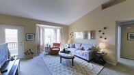 This single-floor residence offers a thoughtfully designed layout that balances comfort and functionality. The living room is a standout space, featuring soaring vaulted ceilings that lend an airy, open feel. A classic bay window floods the room with natural light, complemented by elegant draping curtains. The room is anchored by plush carpeting and a generous seating arrangement, including a tufted ottoman coffee table, a lavender sofa adorned with accent pillows, and a coral wingback armchair. Decorative wall mirrors and sculptural wall accents add visual interest and depth to the neutral, warm-toned walls. Transitioning into the heart of the home, the dining area features rich hardwood flooring and is centered around a round dining table with upholstered chairs beneath a modern pendant light fixture. A framed floral artwork provides an elegant focal point. The adjacent kitchen is open to the dining space and is equipped with warm wood cabinetry, a full suite of appliances including a refrigerator, range, and built-in microwave, and a breakfast bar countertop. Track lighting illuminates the kitchen efficiently, while the overall open sightlines between the living, dining, and kitchen areas create a cohesive and inviting atmosphere throughout.