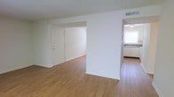 This single-floor space is designed with a straightforward, functional layout that maximizes the use of its open floor plan. The main living area is finished with warm-toned hardwood-style flooring that extends throughout, creating a sense of continuity and cohesion between rooms. Crisp white walls and a textured ceiling reflect natural and artificial light evenly, keeping the interior feeling bright and clean.

A partial wall divides the living area from the adjacent kitchen, offering a degree of separation while maintaining an open, connected feel. The kitchen is visible through the open passageway and features light-colored cabinetry, a sink positioned beneath a window that allows natural light to filter in, and dark countertops that provide contrast to the pale surroundings.

The entry door, positioned within the main living space, is a standard white paneled door with gold hardware, providing direct access to the unit. An overhead HVAC vent is visible near the ceiling, indicating a central air system. The overall ambiance is neutral and move-in ready, offering a clean canvas suitable for a variety of interior styles.