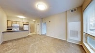 This single-floor residential unit offers a straightforward and functional layout designed with everyday comfort in mind. The open-concept living area is finished with wall-to-wall carpeting in a neutral tone, creating a warm and cohesive feel throughout the main space. Walls are painted in a soft beige, contributing to a calm and understated ambiance that allows for easy personalization.

The kitchen is positioned at one end of the main living area and features light wood cabinetry, a white refrigerator, and a dark countertop, providing a practical workspace with a clean, classic look. A flush-mount ceiling light fixture illuminates the space efficiently, while HVAC vents integrated into the walls and a large wall-mounted panel ensure climate control throughout the unit.

A hallway visible from the main living area leads to additional rooms, offering a sense of separation and privacy within the unit. The entry door, finished in a warm wood tone, provides a welcoming transition from the exterior. Overall, the space is clean, move-in ready, and well-suited for those seeking a no-frills, comfortable living environment with a neutral palette that serves as a versatile backdrop for any furnishing style.