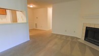 This single-floor residential space offers a straightforward and functional layout designed to maximize comfort and usability. The main living area is generously sized, featuring neutral-toned wall-to-wall carpeting that lends a warm, understated feel to the room. Crisp white walls provide a clean, bright backdrop throughout, offering a versatile canvas for any interior style.

A partial wall with a pass-through opening is visible, suggesting a connected kitchen or dining area that promotes an open, communal flow between spaces. The flush-mount ceiling light provides consistent illumination across the living area, while multiple electrical outlets are conveniently positioned along the walls, reflecting a practical approach to the layout.

A floor vent is integrated into the wall, indicating a built-in climate control system that ensures year-round comfort. The transition between the carpeted living space and what appears to be a hard-surface flooring area beyond adds subtle visual variety while defining separate functional zones within the home. Overall, the space presents a clean, move-in-ready environment with a neutral palette and efficient design, well-suited for a variety of living arrangements.