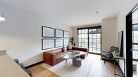 This 1-bedroom model unit showcases a thoughtfully designed, single-floor layout that blends contemporary style with everyday functionality. The open-concept living area is anchored by large, black-framed floor-to-ceiling windows that flood the space with natural light and offer views of the surrounding neighborhood. Light hardwood flooring runs throughout, complementing the clean, neutral color palette of the walls and trim.

The kitchen is a standout feature, equipped with dark cabinetry, light quartz countertops, a white subway tile backsplash, and a full suite of stainless steel appliances. A spacious peninsula with pendant lighting and bar seating creates a natural transition between the kitchen and living areas, making it ideal for both everyday use and entertaining.

The bedroom is a calm, well-lit retreat with multiple black-framed windows that invite natural light from multiple directions. The bathroom features floor-to-ceiling large-format gray tile, a soaking tub with a glass enclosure, and an illuminated mirror, delivering a clean and polished aesthetic. Throughout the unit, consistent design elements — black accents, warm wood tones, and neutral finishes — create a cohesive and modern living environment.
