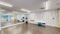 This single-floor fitness studio is thoughtfully designed to provide a clean, functional, and inviting environment for exercise and wellness activities. The space features smooth hardwood-style flooring that extends throughout, offering a durable yet comfortable surface ideal for yoga, stretching, and group fitness classes.

A full-length mirror spans one wall, creating a sense of expanded space while allowing users to monitor their form during workouts. Opposite the mirror, a striking accent wall with a vertical striped pattern in muted tones adds a modern, sophisticated aesthetic to the room. Built-in white shelving lines the lower portion of this wall, providing organized storage for yoga mats and exercise accessories, keeping the floor area open and uncluttered.

The studio is equipped with stability balls displayed on dedicated racks, adding to the room's ready-to-use functionality. Overhead, linear LED lighting fixtures illuminate the space evenly, complemented by natural light filtering in through a window, contributing to a bright and energizing atmosphere.

The overall ambiance is calm yet purposeful — the neutral color palette, clean lines, and deliberate layout make this studio well-suited for a variety of fitness disciplines, from yoga and Pilates to general group exercise and personal training sessions.