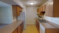 This single-floor residential space is centered around a well-organized galley-style kitchen that serves as its defining feature. The kitchen is designed for efficiency, with warm wood-toned cabinetry lining both sides of the corridor layout, offering generous storage both above and below the countertops. Speckled laminate countertops provide ample workspace for meal preparation, while the white appliance suite — including a refrigerator, electric range, and dishwasher — delivers all the essentials for comfortable daily living. A double-basin sink is conveniently positioned along one wall, complemented by an overhead exhaust vent above the range. The light-toned vinyl flooring runs throughout the kitchen, contributing to a clean and easy-to-maintain finish. A flush-mount ceiling light provides even illumination across the space. The textured ceiling adds a classic residential character, while neutral-toned walls keep the atmosphere open and adaptable. The overall layout is straightforward and practical, making efficient use of the available footprint to deliver a fully equipped and move-in ready living environment.