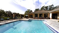 This beautifully designed outdoor amenity space centers around a large, crystal-clear swimming pool that invites relaxation and leisure. The pool area is generously sized, offering ample open water for both swimming and lounging, with a gradual entry point and clean, well-maintained tile work throughout.

Surrounding the pool is a spacious paved sun deck furnished with lounge chairs and seating areas, ideal for outdoor gatherings or quiet relaxation. A shaded umbrella station provides a comfortable retreat from the sun, while additional seating lines the perimeter for socializing.

The focal point of the space is a Mediterranean-inspired clubhouse building featuring a classic terracotta tile roof, warm stucco exterior, and tall decorative columns. Large windows and glass doors allow natural light to flow through the structure, which appears to house an indoor gathering or lounge area. An adjacent pergola-covered patio extends the entertainment space outdoors, seamlessly blending interior and exterior living.

The entire area is framed by mature palm trees and lush greenery, lending a tropical, resort-like ambiance. The surrounding landscape adds both privacy and a sense of natural beauty to the space. Overall, this outdoor amenity area exudes a relaxed yet refined atmosphere, designed for comfort, recreation, and community enjoyment.