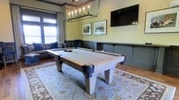 This single-floor space is designed with entertainment and leisure in mind, centered around a full-size billiards table finished in whitewashed wood with dark metal corner accents. The table rests atop a decorative area rug with soft blue and cream tones, adding warmth and texture to the room. Overhead, a striking rectangular candelabra-style chandelier with an industrial black frame provides atmospheric lighting directly above the table.

The walls feature a two-tone treatment — sage green on the upper half and a deep gray wainscoting panel on the lower half — lending the space a refined, classic character. Framed nature artwork adorns the walls alongside a wall-mounted flat-screen television, making the room equally suited for watching sports or socializing.

Natural light filters into the space through windows dressed with neutral drapes, brightening the room without compromising its cozy ambiance. A plush sofa offers comfortable seating along the perimeter, completing the lounge atmosphere. The hardwood flooring visible at the edges of the rug ties the design together with a warm, traditional aesthetic. Overall, this game room strikes a balance between sophisticated interior design and casual, functional recreation space.
