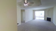 This single-floor residence offers a clean, functional layout designed for comfortable everyday living. The main living area is generously sized, featuring wall-to-wall carpeting that adds warmth underfoot. A decorative tray ceiling draws the eye upward and lends architectural interest to the space, complemented by a ceiling fan with integrated lighting that provides both comfort and illumination. Neutral, light-toned walls throughout create a bright and airy atmosphere, offering a versatile backdrop for a variety of interior styles. A sliding glass door floods the living area with natural light and provides direct access to an outdoor balcony, offering views to the exterior. A hallway visible from the main living space leads to additional rooms, suggesting a thoughtful floor plan that separates living and private areas. The overall ambiance is calm and understated, with a straightforward design that prioritizes openness and livability. The space is well-suited for those seeking a low-maintenance, move-in ready environment with key comfort features already in place.
