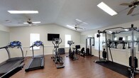 This fitness center offers a clean, well-organized space designed to support a variety of workout routines. The room features vaulted ceilings that lend an open, airy feel, complemented by flush skylight-style panel lighting and ceiling fans that promote air circulation and comfort during exercise. Hardwood-style flooring runs throughout, providing both durability and a polished aesthetic.

The cardio section is equipped with a treadmill, an elliptical trainer, and a recumbent stationary bike, offering options for low- and high-impact cardiovascular workouts. A wall-mounted flat-screen television is positioned for easy viewing during cardio sessions. For strength training, the space includes a multi-function cable machine and a dedicated free-standing strength station, providing a range of resistance training options. An exercise mat is also available for stretching or floor-based workouts.

Natural light filters in through multiple windows along the perimeter, creating a bright and inviting atmosphere. Large mirrors on one wall enhance the sense of space and allow users to monitor their form. The neutral wall tones and uncluttered layout contribute to a focused, functional environment well-suited for daily fitness routines.