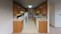This single-floor unit offers a functional and comfortable layout designed for everyday living. The galley-style kitchen is lined on both sides with warm oak cabinetry providing generous storage, complemented by light countertops, a double sink, and white appliances including a range and refrigerator. Patterned vinyl flooring runs the length of the kitchen, adding a classic touch. The open living and dining area is finished with plush neutral carpeting and features a chandelier over the dining space, a wall-mounted air conditioning unit, and a large window that brings in natural light and outdoor views. Baseboard heating is present throughout the unit. The bedrooms are generously sized, each featuring soft carpeting, windows with blinds for natural light and privacy, and baseboard heaters. At least one bedroom includes a built-in closet with a hanging rod for storage. A textured ceiling is consistent throughout the living spaces. The full bathroom is finished with light ceramic tile flooring, a single-sink oak vanity with a large mirror, and a shower/tub combination with a chrome towel bar. The overall palette of warm beige tones and oak wood finishes gives the unit a cohesive and inviting feel throughout.