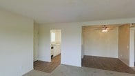 This single-story space presents an open, functional layout that connects multiple living areas with a straightforward design. The main living area features carpeted flooring, which transitions into wood-style hard flooring in the adjoining rooms. A textured popcorn ceiling runs throughout, and a ceiling fan is visible in the adjacent room, providing air circulation.

The kitchen is accessible through an open doorway and is outfitted with white cabinetry, a sink positioned beneath a window that allows natural light to enter, and a dishwasher. The white cabinetry contrasts with the wood-style flooring that continues from the main areas into the kitchen.

A separate room, visible to the right, also features the wood-style flooring and the ceiling fan, suggesting use as a dining area or secondary living space. The walls throughout are painted in a neutral white tone, contributing to a clean and understated aesthetic. The overall layout is practical, with clearly defined zones that are open and connected, making the space feel cohesive and easy to navigate.