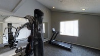 This single-floor fitness space is designed to support a well-rounded workout routine in a focused, functional environment. The room features vaulted ceilings with a textured finish, which add a sense of openness to the otherwise compact layout. Neutral grey walls and dark carpet flooring give the space a clean, modern aesthetic that keeps the focus on the equipment.

The room is equipped with a multi-function cable/weight machine for strength and resistance training, as well as a treadmill for cardiovascular exercise. A wall-mounted mirror is positioned to allow users to monitor their form during workouts. Recessed lighting provides consistent illumination throughout, while a large window fitted with vertical blinds allows natural light to filter into the space, creating a bright and energizing atmosphere.

The overall ambiance is practical and purpose-driven, making it well-suited as a personal or shared fitness facility. The combination of strength and cardio equipment, along with the reflective mirror and natural light, ensures the space is both functional and welcoming for daily exercise routines.