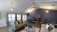 Located in Lancaster, this single-floor space makes a striking first impression with its dramatic vaulted ceilings accented by exposed beam detailing. The open-concept layout connects the living and dining areas in a seamless flow, creating a sense of spaciousness and versatility. A deep navy accent wall serves as the centerpiece of the space, providing a bold contrast against the crisp white ceiling and neutral-toned furnishings. The living area is anchored by a plush gray sofa, a sleek glass coffee table, and a large statement ceiling fan that complements the soaring roofline. Rich, dark hardwood-style flooring runs throughout, adding warmth and cohesion to the overall design. The dining area features a contemporary table set beneath a decorative pendant light, ideal for gatherings. Multiple windows along the far wall allow natural light to fill the space, while French doors offer a bright, welcoming focal point. Thoughtful decorative touches, including tall vases with floral arrangements and layered textiles, contribute to the refined yet comfortable ambiance of this well-appointed Lancaster property.