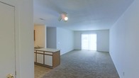 This single-floor space showcases an open-concept design that seamlessly connects the kitchen and living areas. The kitchen features a peninsula with a light-colored countertop, an integrated sink, and warm wood-toned cabinetry below, offering functional prep and storage space. Tile flooring anchors the kitchen zone, transitioning into wall-to-wall carpeting that extends throughout the spacious living area.

The living space is bright and airy, illuminated by a ceiling-mounted light fixture with a track-style design. Neutral gray walls and a smooth ceiling contribute to a clean, understated aesthetic. A large sliding glass door at the far end of the living area floods the space with natural light and provides direct access to an outdoor area, enhancing the sense of openness.

The layout is practical and versatile, with the open sightlines between the kitchen and living room making the space feel expansive. A partial wall separates and defines the kitchen zone without fully enclosing it, maintaining visual connectivity throughout. Overall, the space offers a functional, straightforward design suited for everyday living.